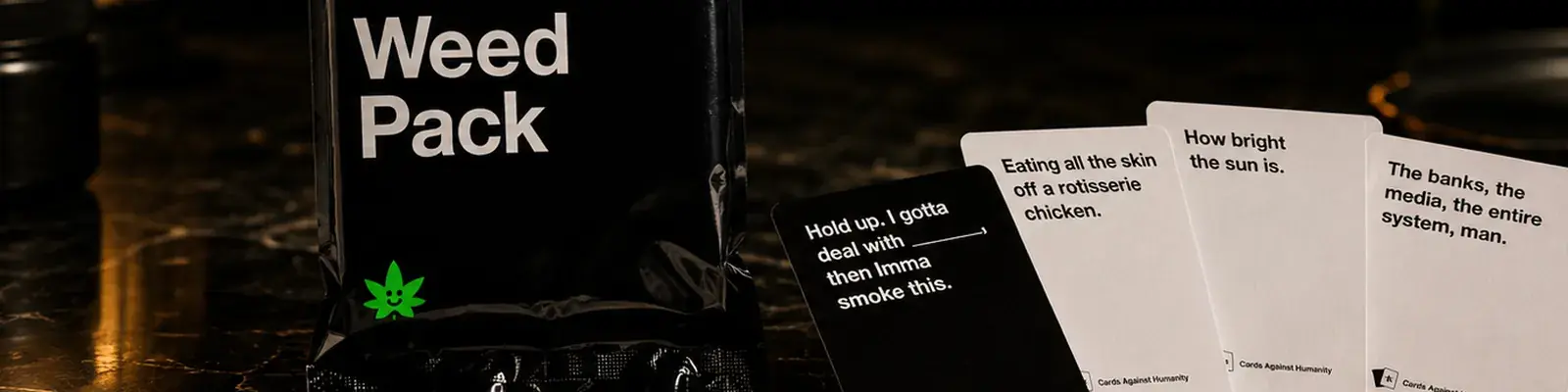 Cards Against Humanity Weed Pack packet beside four fanned cards, wide banner on dark marble