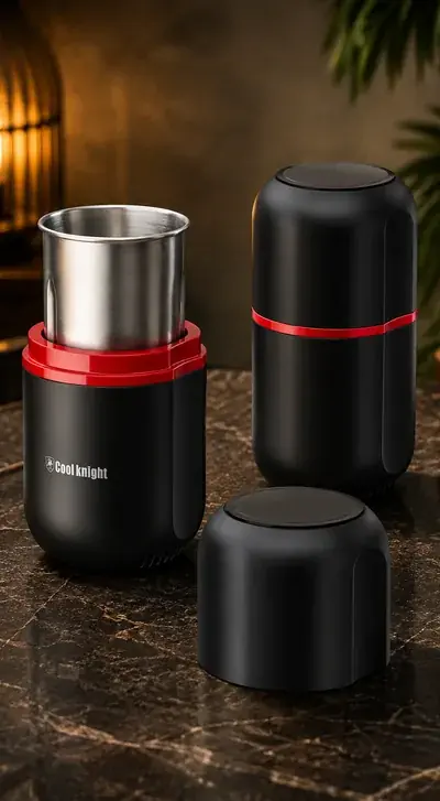 Two black Cool Knight electric herb grinders on dark marble, one open with its stainless grinding bowl lifted clear and one closed, a lid resting in front, warm lamplight behind